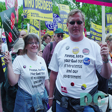 This couple wears coordinating messages on their t-shirts: PLEASE, KEEP YOUR GOD OUT OF MY UTERUS AND OUR GOVERNMENT and PLEASE KEEP GOD OUT OF HER UTERUS AND OUT GOVERNMENT.  In addition they wear many of the stickers and buttons distributed at the march: MARCH FOR WOMENS LIVES, BUCK FUSH, NARAL, STAND UP FOR CHOICE AND KERRY FOR PRESIDENT.
