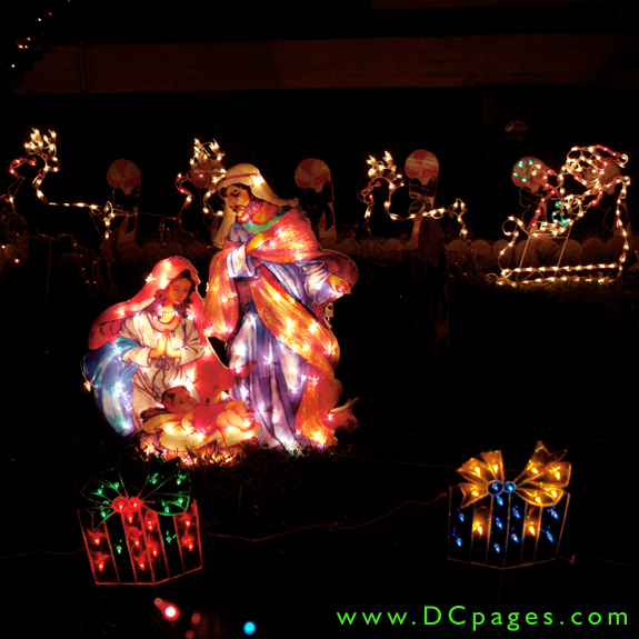 Baby Jesus, Mary, and Joseph are surrounded by lit Christmas presents. A lit Santa and his reindeer are in the background.