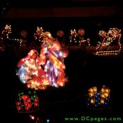 Baby Jesus, Mary, and Joseph are surrounded by lit Christmas presents. A lit Santa and his reindeer are in the background.