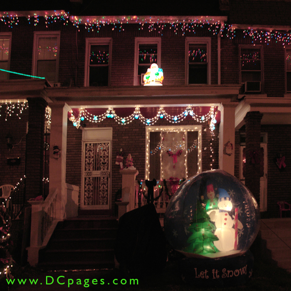 A brightly lit Santa Claus stands on top of a porch wraped in a strand of classic blue and white lights. A red ribbon, snownen, and garland can be seen. A giant inflatable "Let it Snow" globe is in the front of yard.