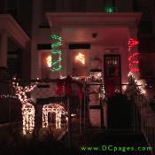 Two glowing reindeer graze inside this yard surrounded in red bows. White and green, and red strings of holiday lights are on display. Two more reindeer can be seen on both windows.