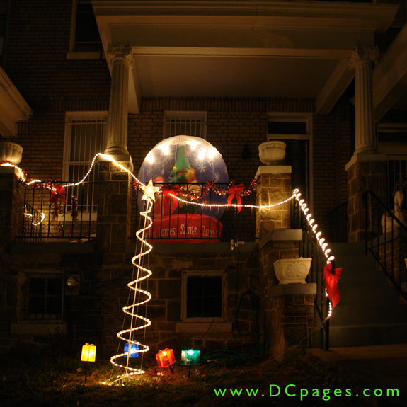A giant "Here Comes Santa Claus" globe is on the front porch of this home. A spiral white tree surrounded by yellow, blue, orange, and green presents is on display in the front yard.
