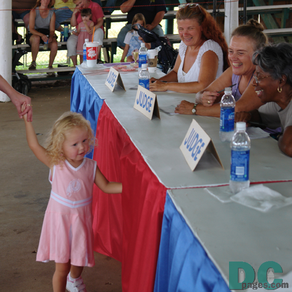 A blond haired baby girl in a pink Washington Redskins dress walks past the judges. 