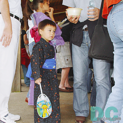 Among tall adults, a little boy was standing with a hand fan next to his sister. 