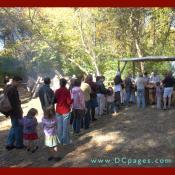 A line of adults and children eagerly wait for some tasty colonial food. 