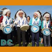 Several tribal men in feathered headdress perform on colorful drums.
