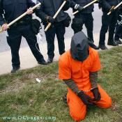 Kneeling Guantanamo Bay detainee actor is wearing an orange jumpsuit, a black bag over his head, with wrists handcuffed together. Riot police holding batons in the background.
